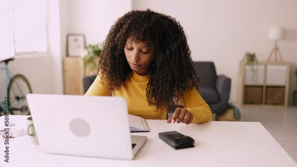 Smiling young african american woman working with laptop computer at home office - Female entrepreneur using calculator while taking notes at workplace Smiling young african american woman working with laptop computer at home office - Female entrepreneur using calculator while taking notes at workplace