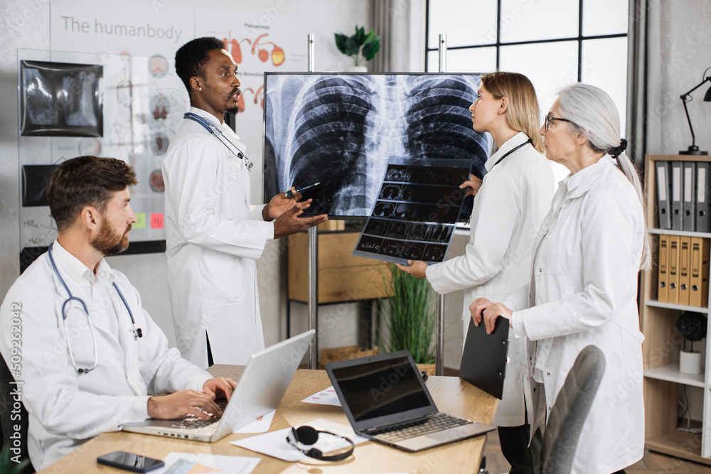 Caucasian female doctor holding x ray scan and talking with multiethnic colleagues while standing near monitor with patient result. Foure medical therapists cooperating for making diagnosis. Caucasian female doctor holding x ray scan and talking with multiethnic colleagues while standing near monitor with patient result. Foure medical therapists cooperating for making diagnosis.
