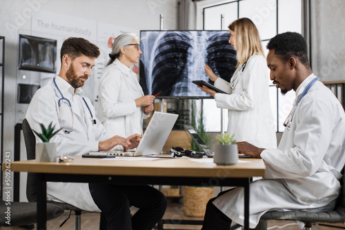 Team of two multi ethnic male medical workers typing on wireless laptops while female colleagues discussing tomography results and making diagnosis. Healthcare and modern technology concept.