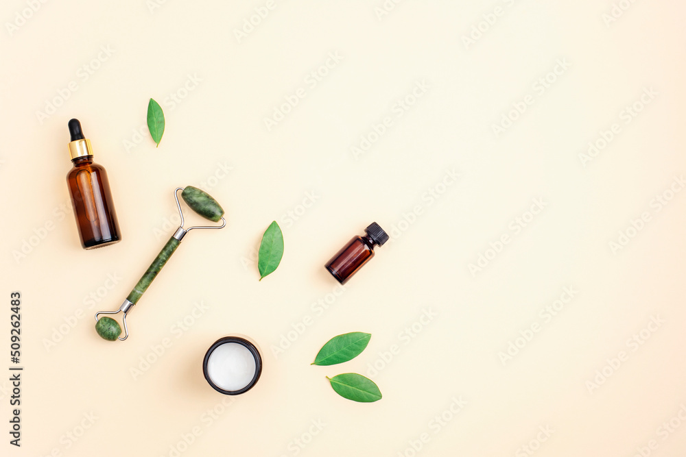 Cream in a jar, serum bottle and jade facial roller on neutral beige background with green leaves, top view. Flat lay, copy space Cream in a jar, serum bottle and jade facial roller on neutral beige background with green leaves, top view. Flat lay, copy space