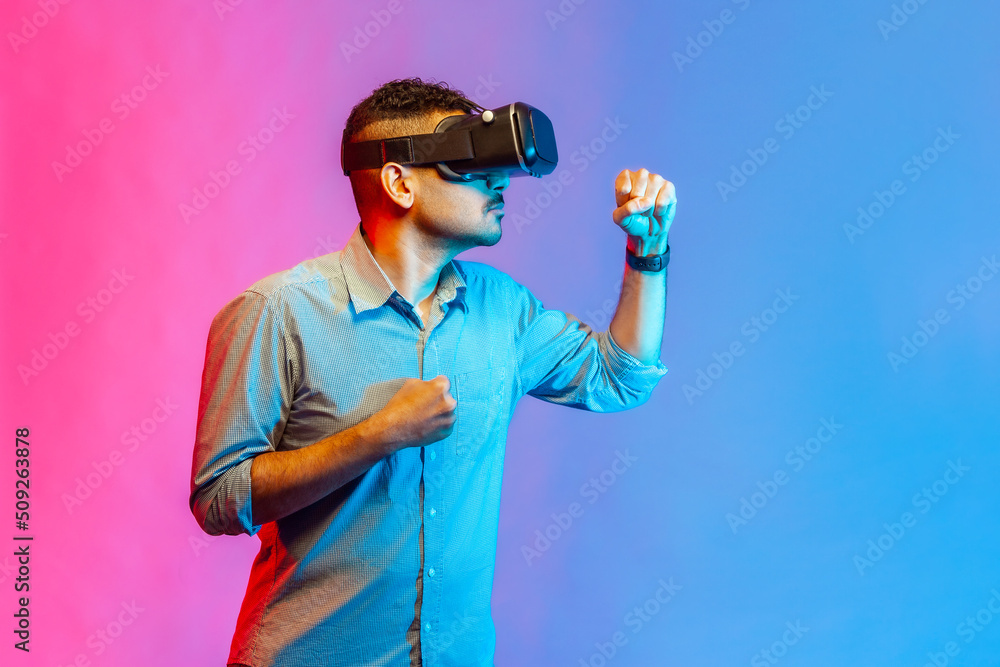 Man in shirt in virtual reality glasses on head playing fighting game, holding clenched fists up ready to boxing, using VR headset tech. Indoor studio shot isolated on colorful neon light background.