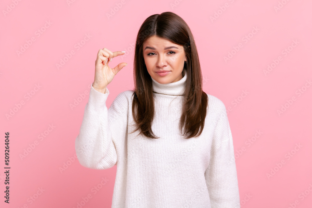 Young adult woman showing a little bit gesture, inch or centimeter, disappointed with minimum size, wearing white casual style sweater. Indoor studio shot isolated on pink background. Young adult woman showing a little bit gesture, inch or centimeter, disappointed with minimum size, wearing white casual style sweater. Indoor studio shot isolated on pink background.