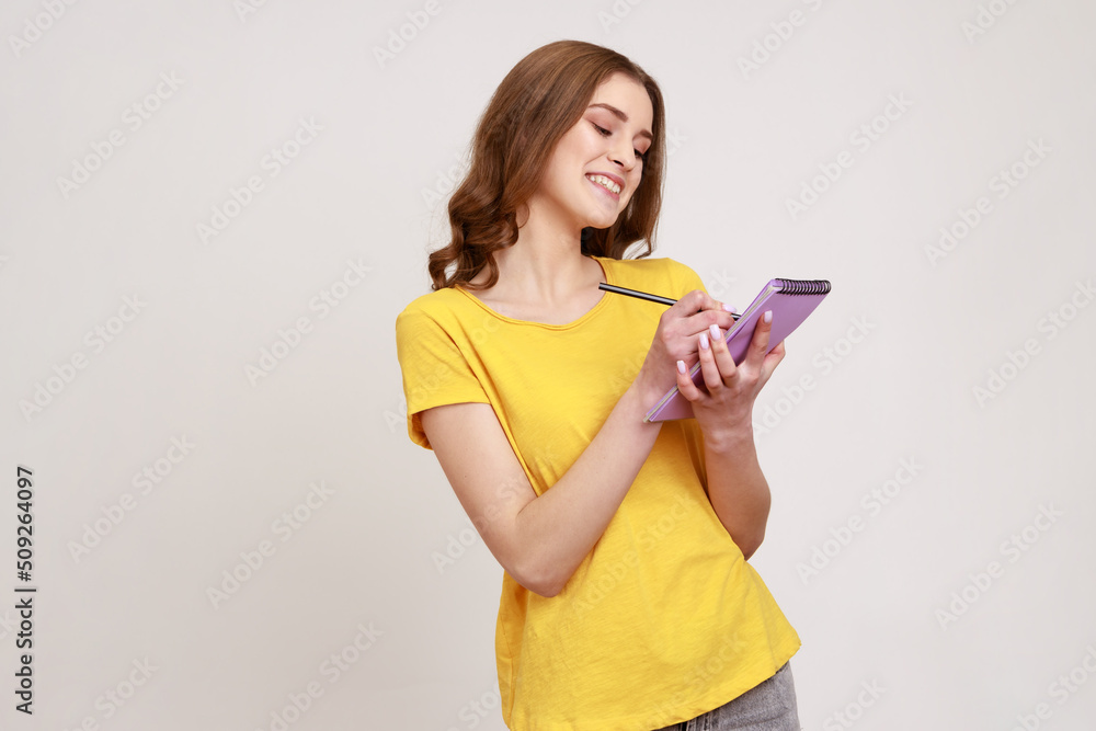 Portrait of glad curly haired teenage girl in yellow casual style T-shirt writing in notebook, filling diary or schedule, smiling sincerely. Indoor studio shot isolated on gray background. Portrait of glad curly haired teenage girl in yellow casual style T-shirt writing in notebook, filling diary or schedule, smiling sincerely. Indoor studio shot isolated on gray background.