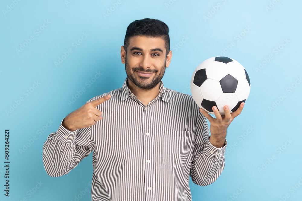 Portrait of handsome businessman pointing finger at soccer ball on his hand with smiling positive expression, wearing striped shirt. Indoor studio shot isolated on blue background. Portrait of handsome businessman pointing finger at soccer ball on his hand with smiling positive expression, wearing striped shirt. Indoor studio shot isolated on blue background.
