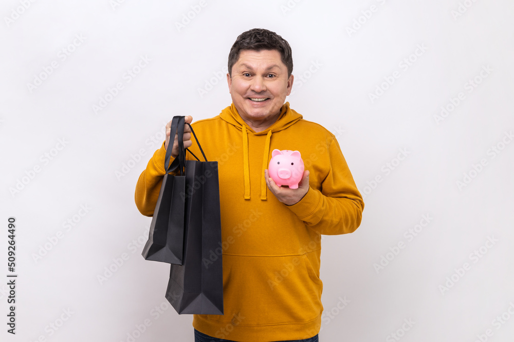 Middle aged man holding shopping bags and piggy bank, saving money, cashback, looking at camera with positive expression, wearing urban style hoodie. Indoor studio shot isolated on white background. Middle aged man holding shopping bags and piggy bank, saving money, cashback, looking at camera with positive expression, wearing urban style hoodie. Indoor studio shot isolated on white background.