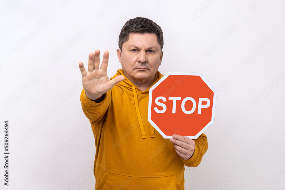 Concentrated serious man looking angrily showing stop gesture, holding road traffic sign, warning of ban, forbidden access, wearing urban style hoodie. Indoor studio shot isolated on white background. Concentrated serious man looking angrily showing stop gesture, holding road traffic sign, warning of ban, forbidden access, wearing urban style hoodie. Indoor studio shot isolated on white background.