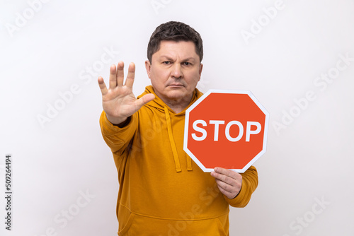 Concentrated serious man looking angrily showing stop gesture, holding road traffic sign, warning of ban, forbidden access, wearing urban style hoodie. Indoor studio shot isolated on white background. © khosrork