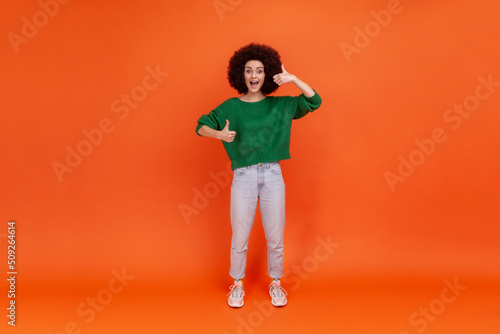 Full length portrait of happy friendly woman with Afro hairstyle wearing green casual style sweater standing with thumbs up, expressing positive. Indoor studio shot isolated on orange background.
