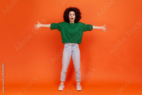 Full length portrait of woman with Afro hairstyle wearing green casual style sweater standing with raised arms, looking with shocked expression. Indoor studio shot isolated on orange background.