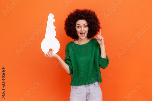 Excited woman with Afro hairstyle wearing green casual style sweater holding paper key in hand and raising finger up, having idea. Indoor studio shot isolated on orange background.