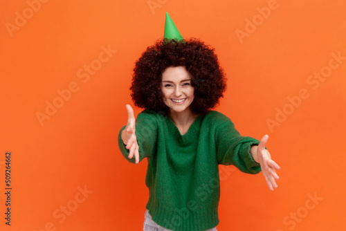 Happy satisfied woman with Afro hairstyle wearing green casual style sweater and cone, celebrating birthday party, inviting guests. Indoor studio shot isolated on orange background.