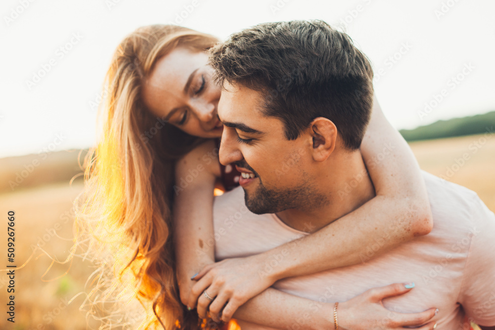 Fototapeta premium Portrait of smiling handsome man giving his ginger girlfriend a piggy ride back in wheat field. Beautiful portrait. Leisure activities. Summer activities