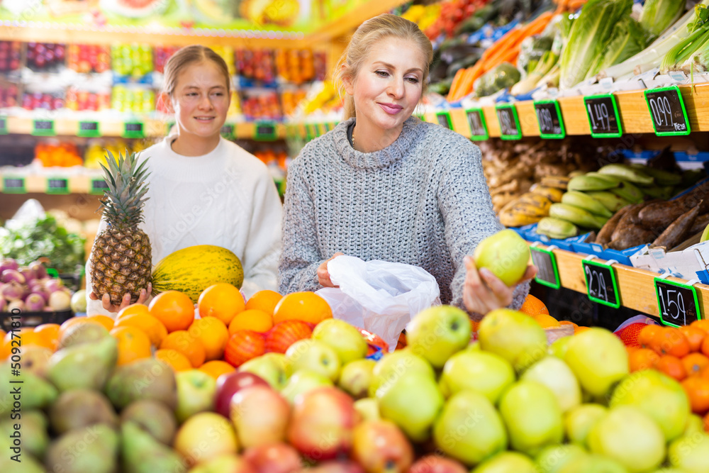 Portrait of cheerful friendly teenage girl and her mother during family shopping in fruit store..