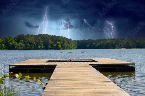 a long brown wooden dock over vast blue rippling lake water surrounded by lush green trees with people in colorful kayaks on the water and powerful clouds with lightning at Cauble Park in Acworth photo