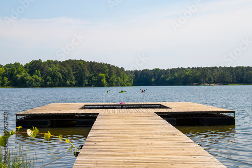 a long brown wooden dock over vast blue rippling lake water surrounded by lush green trees with people in colorful kayaks on the water and blue sky at Cauble Park in in Acworth Georgia USA photo