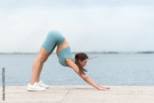A beautiful girl is doing yoga by the river . A young woman in sports clothes near the water