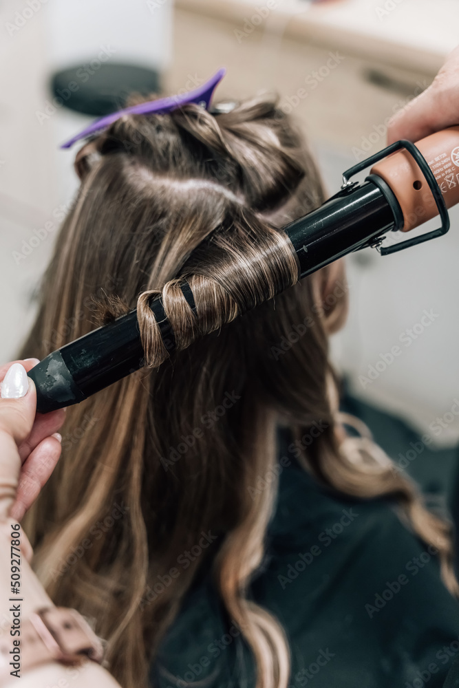 Naklejka premium Female hands of a hair master. Close-up of blond hair on a curling iron. Curling curls.