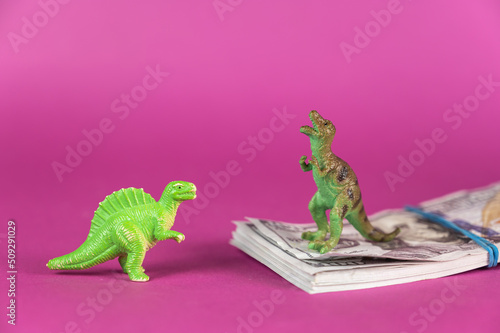 Dinosaur miniatures and a wad of hundred-dollar bills against a purple background. A stack of money tied with a rubber band. Green figures of animals of prey standing on their hind legs. © Mikhail