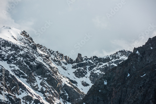 Dramatic landscape with sunlit high snowy mountain with peaked top in cloudy sky. White snow on black sharp rocks with golden shine. Awesome snow mountain peak with gold sunlight in changeable weather