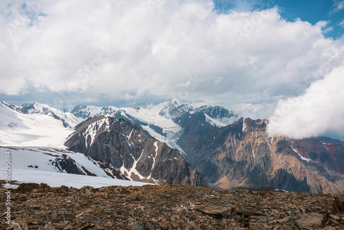 Dramatic landscape with large glacier and high snow mountains in cloudy sky. Wonderful view from stone hill with snow to glacier and snowy mountain peak and low clouds. Top view to majestic mountains.