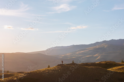 Tourist with camera on sunlit gold grassy hill with dirt road with view to high mountains. Beautiful sunny landscape with man in mountains. Colorful scenery with traveler on hill in golden sunlight.