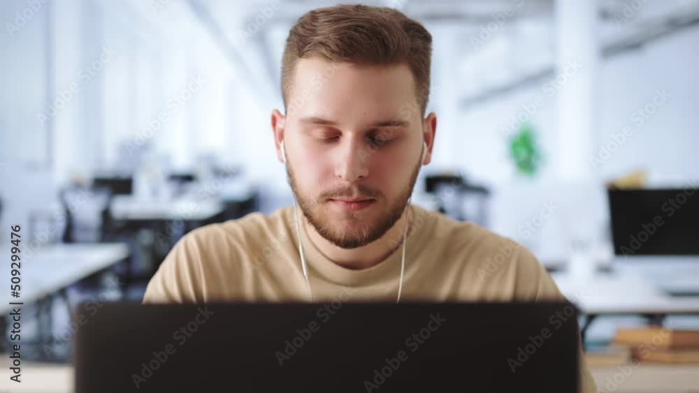 Bearded young man typing on wireless laptop in empty office room. Focused entrepreneur in modern earphones working on new project. People and technology concept. Bearded young man typing on wireless laptop in empty office room. Focused entrepreneur in modern earphones working on new project. People and technology concept.