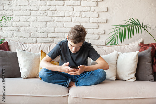 A teenage boy sits on the couch with a smartphone in his hands. Digital addiction.