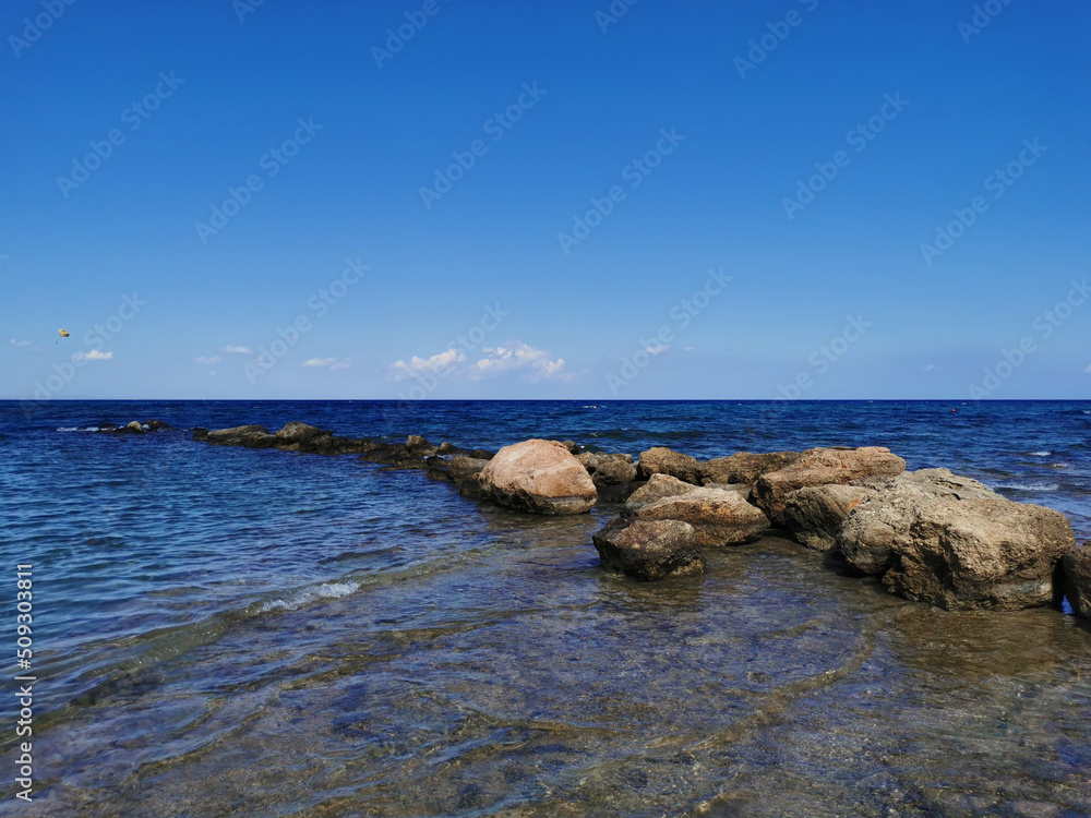 The coast of the Mediterranean Sea, waves, clear water, a stone ridge against a blue sky with clouds.