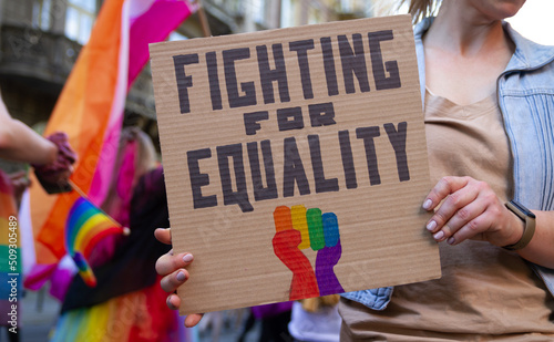 Woman holding placard sign Fighting for Equality with rainbow flag fist, during LGBT Pride Parade. Crowd of people at equality march to support and celebrate LGBT+, LGBTQ gay and lesbian community.