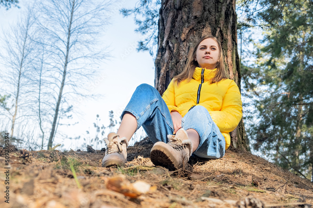 Portrait of a happy smiling young girl sitting in the woods under a tree. Mental health. The woman wears a bright yellow jacket. Time for yourself, reboot, meditation. Portrait of a happy smiling young girl sitting in the woods under a tree. Mental health. The woman wears a bright yellow jacket. Time for yourself, reboot, meditation.
