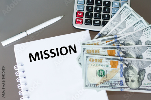 Text MISSION on a notepad on a table next to a pen, calculator and money