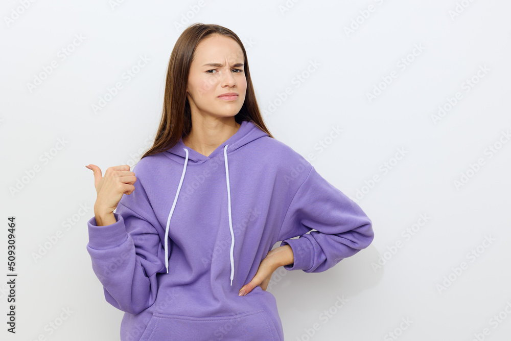 an emotional woman stands on a gray background in a purple tracksuit with a dissatisfied thumb pointing behind her back slightly wrinkling her forehead an emotional woman stands on a gray background in a purple tracksuit with a dissatisfied thumb pointing behind her back slightly wrinkling her forehead