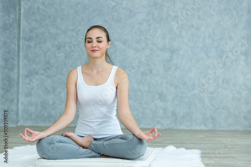 A beautiful woman with neat ponytail hairstyle is practicing yoga and meditates in the lotus position while sitting on the soft white mattress in the yoga class background. Heathy lifestyle concept.
