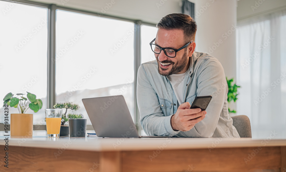 Smiling businessman looking at laptop while holding cellphone at desk in office