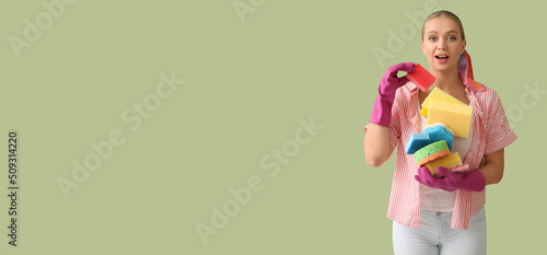 Surprised young woman with many cleaning sponges on green background with space for text