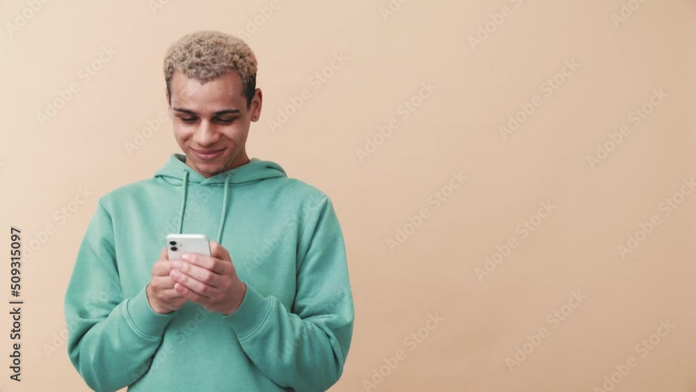Cheerful curly-haired man wearing blue hoodie typing by phone in the beige studio Cheerful curly-haired man wearing blue hoodie typing by phone in the beige studio