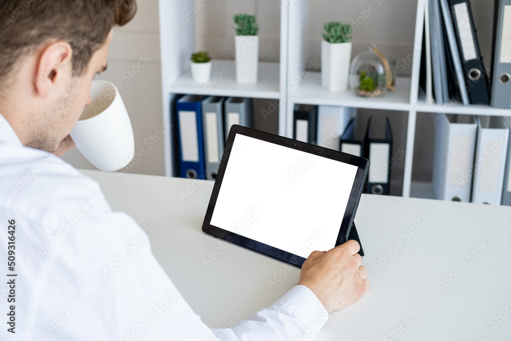 Fototapeta premium Office work. Computer mockup. Digital technology. Unrecognizable man looking tablet computer with blank screen sitting desk drinking coffee in light room interior.