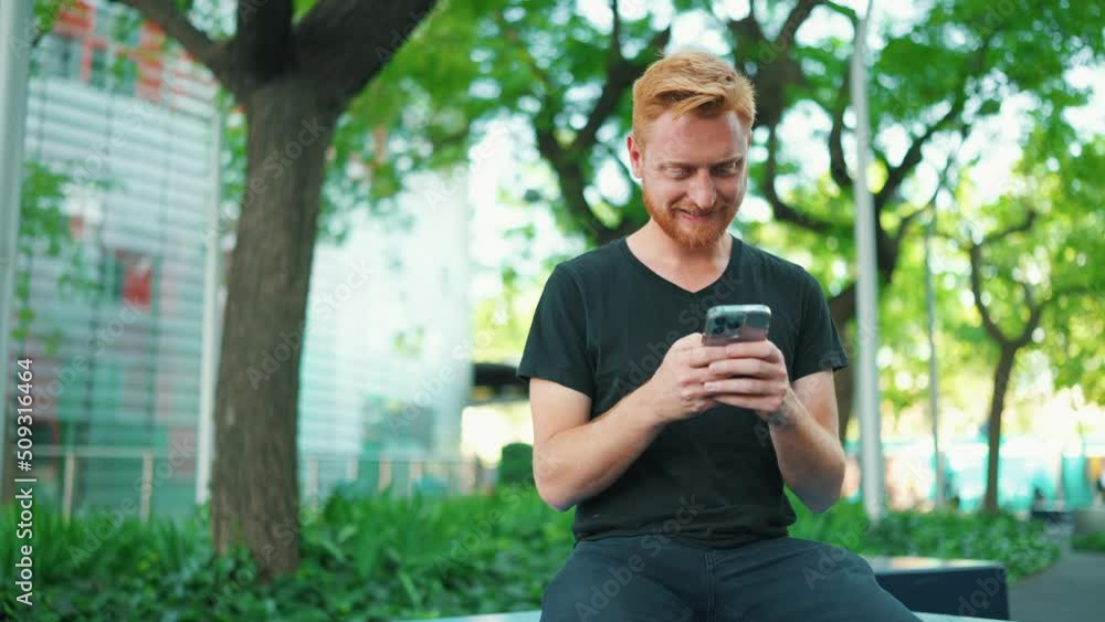 Smiling red haired man texting by smartphone while sitting on the bench in the park Smiling red haired man texting by smartphone while sitting on the bench in the park