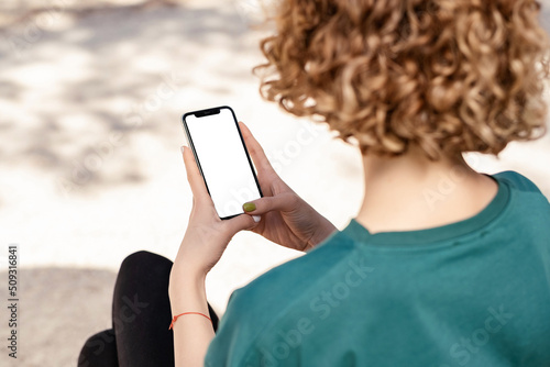 Beautiful redhead woman wearing sportive clothes standing on city park, outdoor holding phone touching finger mockup white blank display, mobile application tech concept, over shoulder closeup view.