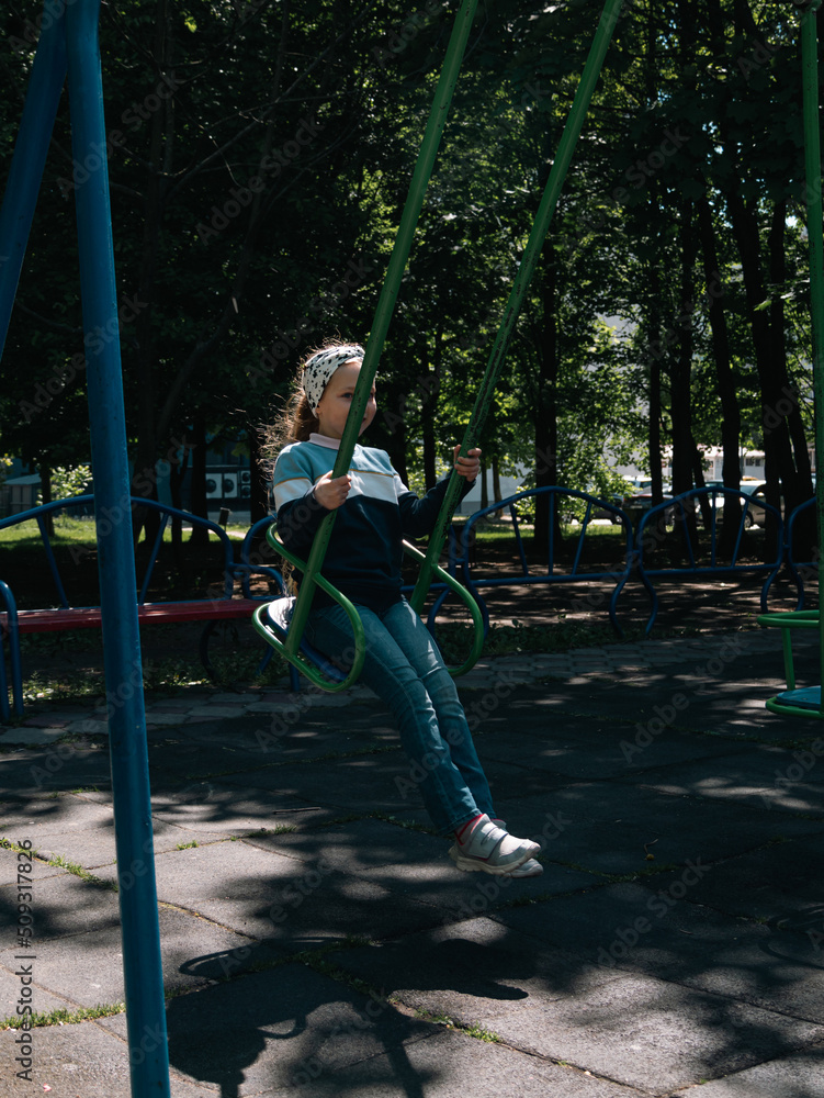 Obraz premium Happy child on a swing in sping park on vacation. Cute little girl with long blond hair swinging on a playground alone in tree shadow. Joy, fun, summer activities on fresh air.