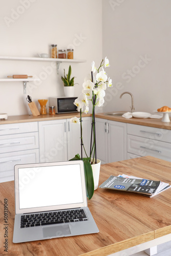 Modern laptop, orchid flower and magazines on dining table in kitchen