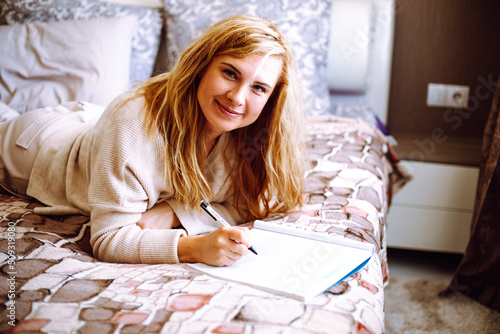 Smiling blonde woman in home clothes lying on bed and writing with pen in notebook closeup. Young happy lady relaxing at home in cozy bedroom in morning. Working and useful activity concept