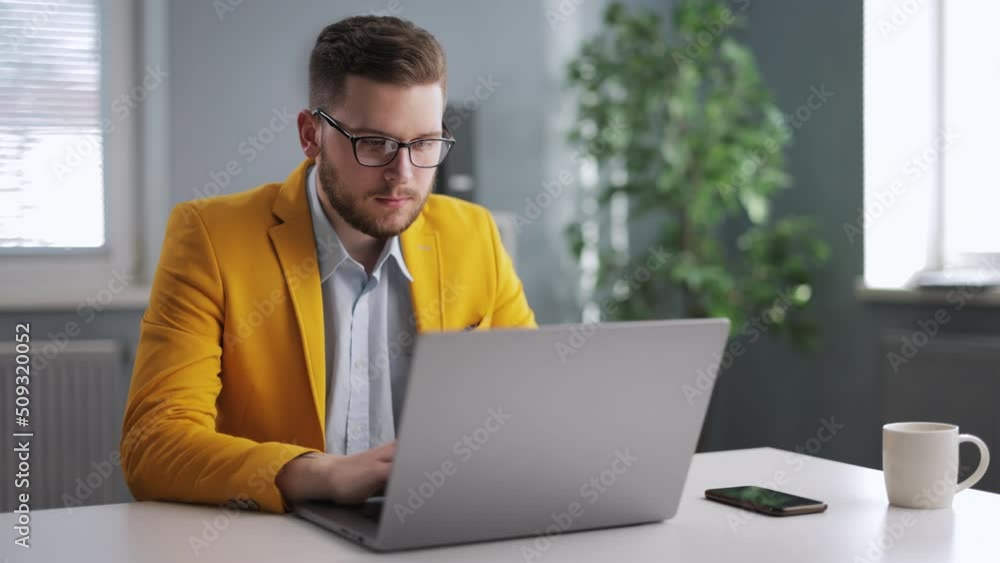 Handsome young man in glasses and yellow jacket working on wireless laptop while sitting at office. Caucasian bearded male sitting at desk and looking on computer screen. Handsome young man in glasses and yellow jacket working on wireless laptop while sitting at office. Caucasian bearded male sitting at desk and looking on computer screen.