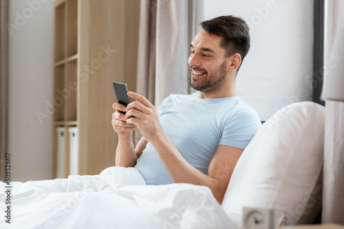 people, bedtime and rest concept - happy smiling man with smartphone lying in bed at home