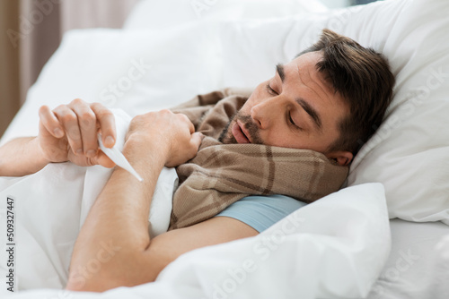 people, health and fever concept - sick man in scarf measuring temperature by thermometer lying in bed at home