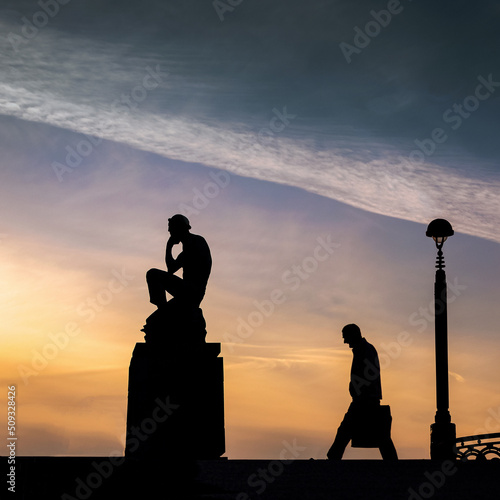 An business man walks and thinks in the sunset next to an old man meditative sculpture