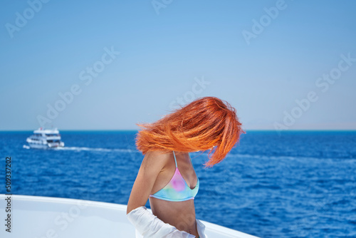 Portrait of red hair girl posing on the edge of white yacht, arms outstretched in different directions, enjoying the flow of wind. Concept of freedom and summer vacation. 