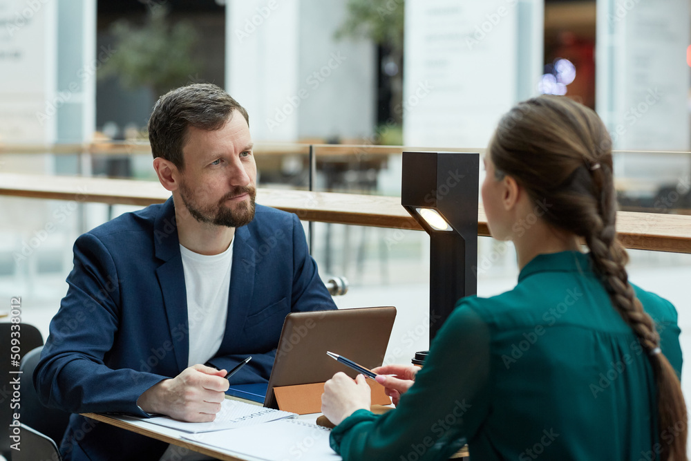 Portrait of bearded businessman listening to young woman during meeting at cafe table with documents - obrazy, fototapety, plakaty 
