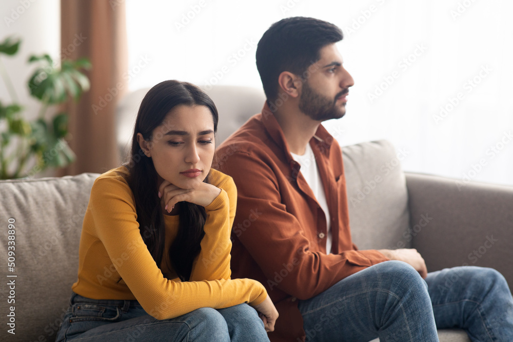 Obraz premium Unhappy Arabic Couple Sitting On Couch Indoor, Having Relationship Problems