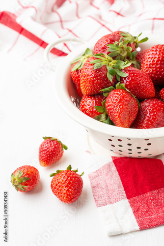 Aerial view of strawberries in a white colander on a white table with a red kitchen cloth, white background, vertical, with copy space